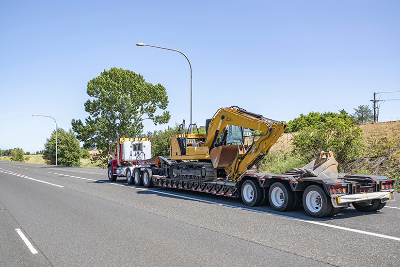 Industrial standard professional red day cab big rig heavy duty semi truck tractor transporting fastened construction equipment on step down semi trailer driving on the wide highway road